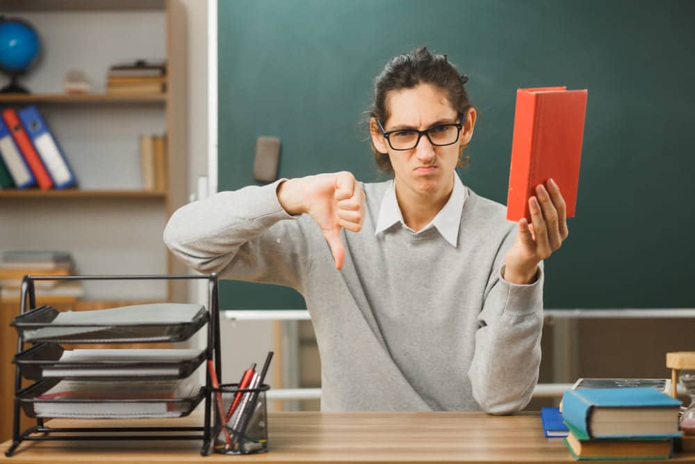 unpleased showing thumbs down young male teacher holding book sitting desk with school tools classroom 2 Clases de alemán online